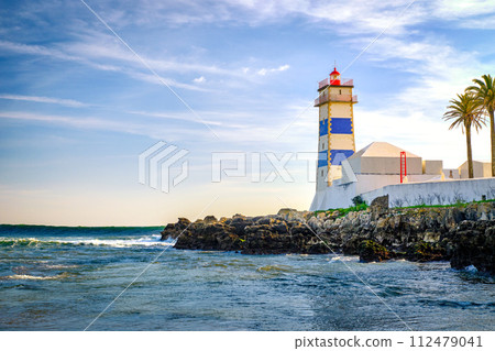 Beautiful evening, on sea tidal waves, rocky shores and Santa Marta lighthouse in Cascais, Portugal. Blue sky, clouds, sunlight, low sun, calm ocean waters, local landmark, safety and navigation tower 112479041