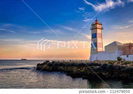 Dramatic sunset on sea shore and Santa Marta lighthouse in Cascais, Portugal. Colorful sky, clouds, sunlight, low sun, calm ocean waters, local landmark, safety and navigation tower, rocky shore. Dramatic sunset on sea shore and Santa Marta lighthouse in Cascais, Portugal. Colorful sky, clouds, sunlight, low sun, calm ocean waters, local landmark, safety and navigation tower, rocky shore. 112479050