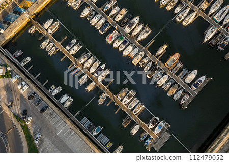 Aerial top down view of a row of yachts, sailboats and jetties in marina with moored sailboats. Low sun, sunshine, drone stay and ascend revealing more vessels. 112479052