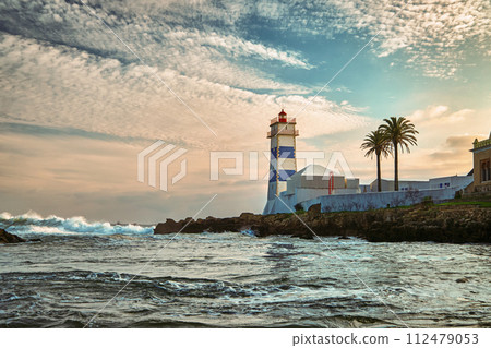 Beautiful sunset landscape on sea shore and Santa Marta lighthouse in Cascais, Portugal. Colorful sky, clouds, sunlight, low sun, calm ocean waters, local landmark, safety and navigation tower, rocky Beautiful sunset landscape on sea shore and Santa Marta lighthouse in Cascais, Portugal. Colorful sky, clouds, sunlight, low sun, calm ocean waters, local landmark, safety and navigation tower, rocky 112479053