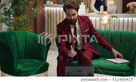 White collar worker attends videocall meeting, sitting in lounge area at modern hotel and talking to executive board and shareholders on teleconference. Businessman in suit presenting new ideas. 112480417