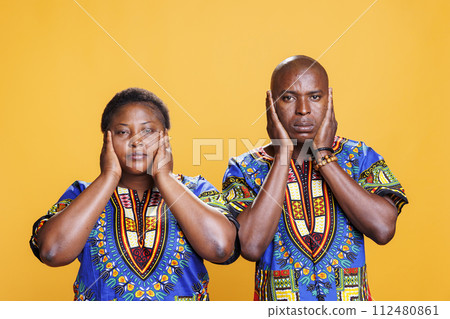 African american couple covering ears with hands, showing hear no evil three wise monkeys portrait. Man and woman ignoring loud noise and looking at camera on studio background 112480861