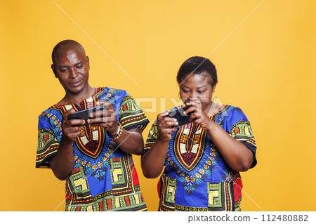 Cheerful african american wife and husband pair enjoying mobile game competition. Smiling couple playing entertaining game on smartphones, sharing fun leisure activity together 112480882
