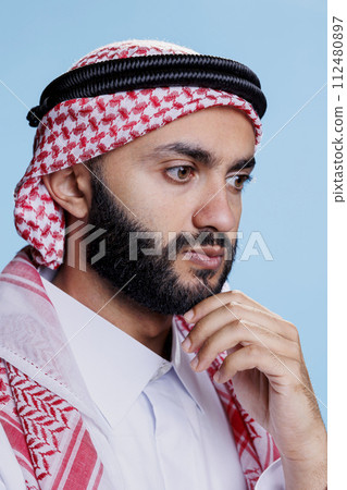 Muslim man in traditional headdress thinking and considering solution. Arab person wearing ghutra headscarf rubbing chin and looking away with concentrated expression closeup Muslim man in traditional headdress thinking and considering solution. Arab person wearing ghutra headscarf rubbing chin and looking away with concentrated expression closeup 112480897
