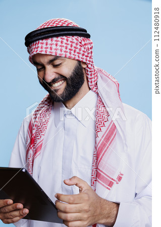Arab man laughing while enjoying internet entertainment activity on digital tablet. Smiling muslim person having fun while chatting online and scrolling social media on gadget touchscreen Arab man laughing while enjoying internet entertainment activity on digital tablet. Smiling muslim person having fun while chatting online and scrolling social media on gadget touchscreen 112480918