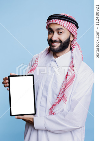 Smiling arab dressed in traditional attire holding digital tablet with white blank screen mockup and looking at camera. Muslim man promoting portable gadget empty touchscreen for application ads Smiling arab dressed in traditional attire holding digital tablet with white blank screen mockup and looking at camera. Muslim man promoting portable gadget empty touchscreen for application ads 112480921