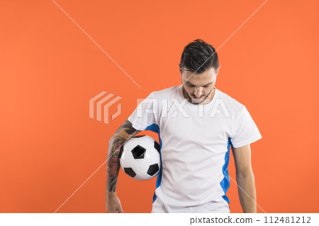 Soccer player looking down and holding a ball with one arm isolated on orange background 112481212