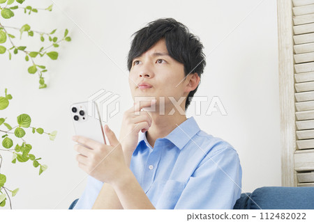 Young man using smartphone while thinking in living room 112482022