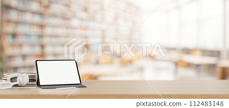 A tablet mockup, headphones, and books on a wooden desk with a blurred background of a library. 112483148