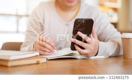 A cropped shot of a woman using her phone while working on her tasks at a table in a coffee shop. A cropped shot of a woman using her phone while working on her tasks at a table in a coffee shop. 112483275