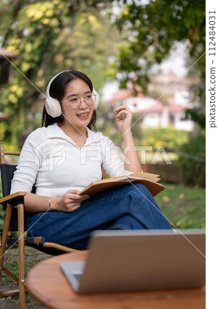 Happy Asian woman is listening to music, working or writing her diary while sitting in her backyard. 112484031