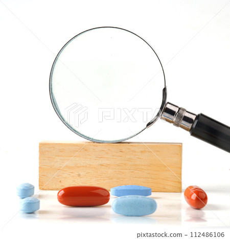 Magnifying glass on a wooden bar in close-up with pills in the foreground. A place to c 112486106