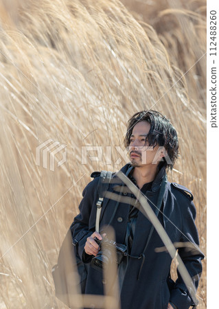A man in his 30s holding a camera in front of pampas grass A man in his 30s holding a camera in front of pampas grass 112488260