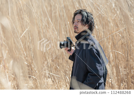 A man in his 30s holding a camera in front of pampas grass 112488268