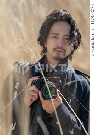 A man in his 30s holding a camera in front of pampas grass A man in his 30s holding a camera in front of pampas grass 112488278