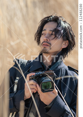 A man in his 30s holding a camera in front of pampas grass A man in his 30s holding a camera in front of pampas grass 112488280