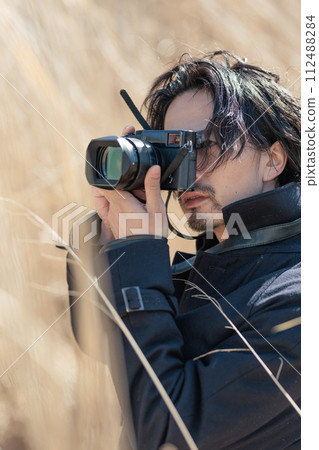 A man in his 30s holding a camera in front of pampas grass A man in his 30s holding a camera in front of pampas grass 112488284