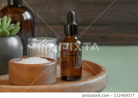 Natural cosmetics advertising scene. Two amber bottles is on a wooden plate with sea bath salt and moisturizer. Blurred wood background. Essential oils. Place for text. SPA massaging concept. Health 112489377