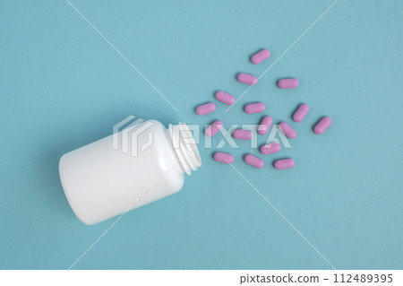 Oval purple pills spilled out of a white jar isolated on a blue color studio background. Mock-up. Banner. Copy space. Place for logo, advertising. Medicine, treatment, capsule for curing disease 112489395