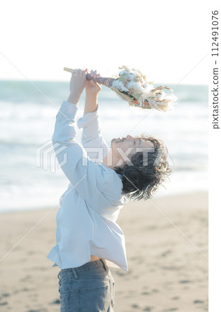 A man in his 30s wearing a white shirt standing by the seaside 112491076