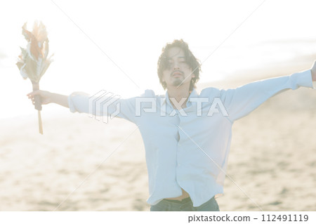A man in his 30s wearing a white shirt standing by the seaside 112491119