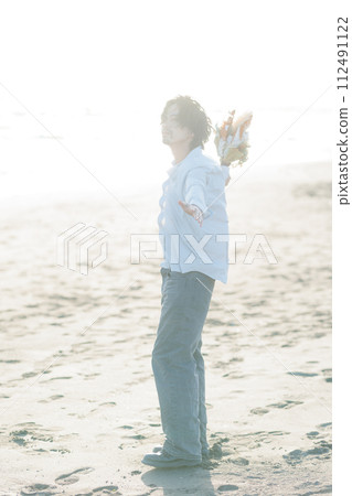 A man in his 30s wearing a white shirt standing by the seaside 112491122