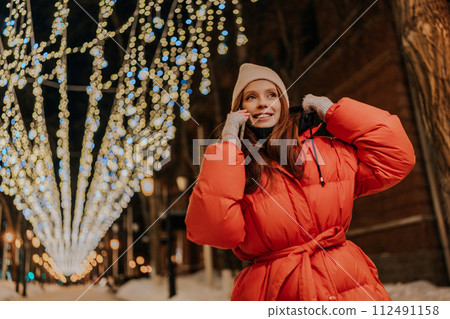 Low-angle view of happy young woman in hat and winter jacket talking using mobile phone standing on dark snow city street, on blurred background of hinged bright festive colorful illumination. Low-angle view of happy young woman in hat and winter jacket talking using mobile phone standing on dark snow city street, on blurred background of hinged bright festive colorful illumination. 112491158