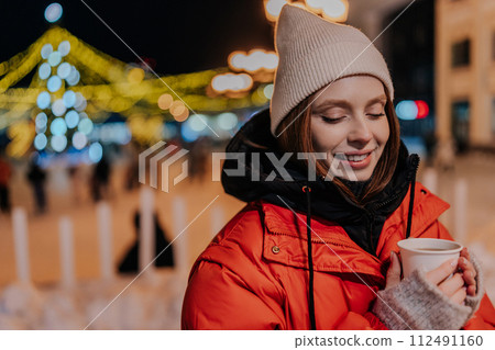 Portrait of charming young woman wearing warm winter clothes enjoying drinking takeaway cup of coffee outdoors on cold street at night with festive bright illumination. Pretty female walking on park Portrait of charming young woman wearing warm winter clothes enjoying drinking takeaway cup of coffee outdoors on cold street at night with festive bright illumination. Pretty female walking on park 112491160