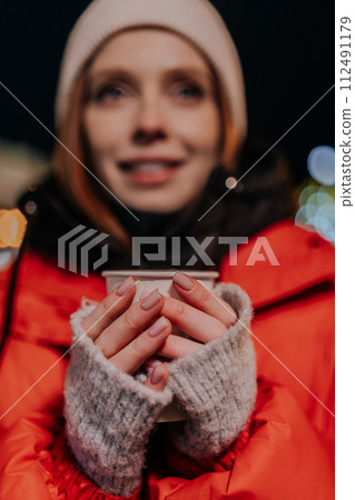 Closeup vertical selective focus portrait of charming young woman wearing warm winter clothes enjoying drinking takeaway cup of coffee outdoors on cold street at night. Pretty female walking on park 112491179