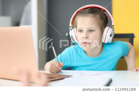 Little girl schoolgirl in headphones sitting in front of laptop at home Little girl schoolgirl in headphones sitting in front of laptop at home 112491189