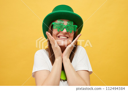 Irish hat and clover celebration. Cultural traditions in Ireland. Cute charming Caucasian woman wearing green leprechaun hat and clover glasses standing isolated over yellow background 112494384
