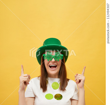 National holiday event. St. Patrick's Day mythology. Happy Caucasian woman wearing green leprechaun hat and clover glasses standing isolated over yellow background pointing up at advertisement area National holiday event. St. Patrick's Day mythology. Happy Caucasian woman wearing green leprechaun hat and clover glasses standing isolated over yellow background pointing up at advertisement area 112494387