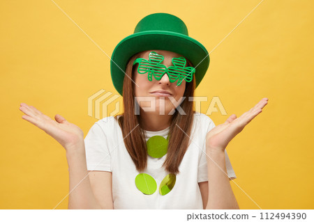 Fun and festive Irish party. Greetings for Irish celebrations. Uncertain woman wearing festive green hat and shamrock glasses standing isolated over yellow background shrugging shoulders Fun and festive Irish party. Greetings for Irish celebrations. Uncertain woman wearing festive green hat and shamrock glasses standing isolated over yellow background shrugging shoulders 112494390