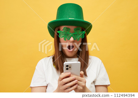 Mythology and tradition intertwined in event. Shocked Caucasian woman wearing green leprechaun hat and clover glasses standing isolated over yellow background scrolling online reading breaking news 112494394