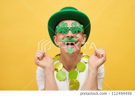Mythology and fun celebration. Extremely happy Caucasian man wearing green leprechaun hat and clover glasses standing isolated over yellow background clenched fists with happy face Mythology and fun celebration. Extremely happy Caucasian man wearing green leprechaun hat and clover glasses standing isolated over yellow background clenched fists with happy face 112494498