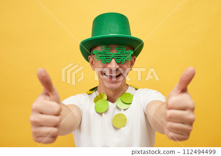 National holiday fun. Smiling man wearing festive green hat and shamrock glasses standing isolated over yellow background showing thumb up like gesture National holiday fun. Smiling man wearing festive green hat and shamrock glasses standing isolated over yellow background showing thumb up like gesture 112494499