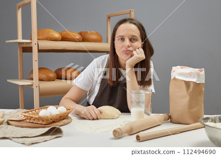 Sad unhappy woman wearing brown apron kneading yeast dough with hand to baking bread while sitting at table on table in her bakery looking at camera with unhappy bored face Sad unhappy woman wearing brown apron kneading yeast dough with hand to baking bread while sitting at table on table in her bakery looking at camera with unhappy bored face 112494690