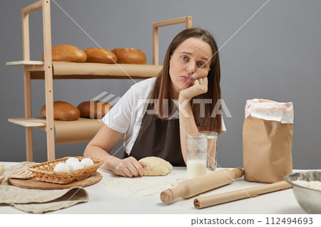Tired exhausted upset woman wearing brown apron kneading yeast dough with hand on table looking at camera preparing dough for bread baking Tired exhausted upset woman wearing brown apron kneading yeast dough with hand on table looking at camera preparing dough for bread baking 112494693