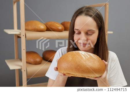 Dark haired smiling satisfied woman holding fresh bread enjoying baking homemade pastry cooking with love and pleasure in her bakehouse Dark haired smiling satisfied woman holding fresh bread enjoying baking homemade pastry cooking with love and pleasure in her bakehouse 112494715