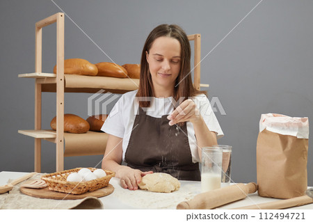 Attractive busy woman wearing brown apron kneading dough sprinkling with flour preparing for baking bread or pie at bakery workplace Attractive busy woman wearing brown apron kneading dough sprinkling with flour preparing for baking bread or pie at bakery workplace 112494721