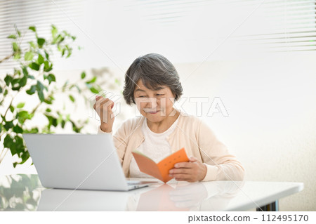 A senior woman using a computer while looking at her pension notebook, ending her life 112495170