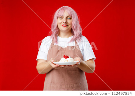 Baker in apron white t-shirt holding in hand cake on plate isolated on red color background in studio. Food or holiday concept Baker in apron white t-shirt holding in hand cake on plate isolated on red color background in studio. Food or holiday concept 112497834