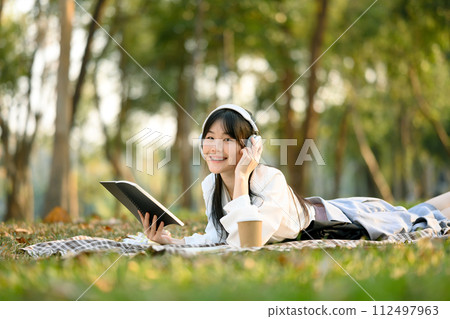 Cheerful young Asian girl in headphones lying on picnic blanket and reading book Cheerful young Asian girl in headphones lying on picnic blanket and reading book 112497963