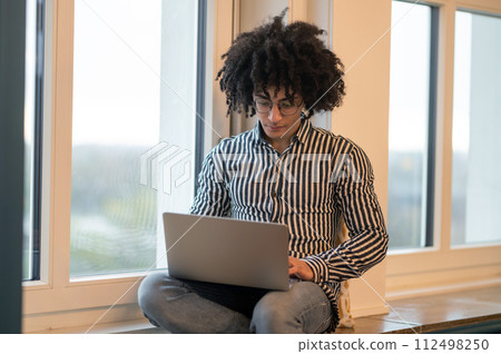 Curly-haired man sitting on a window-sill and working on laptop Curly-haired man sitting on a window-sill and working on laptop 112498250
