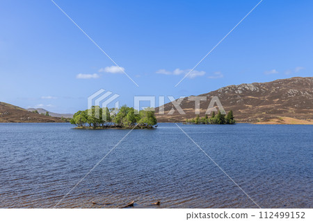 In Scotland's heart, a serene loch cradles a lush island with two red deer, framed by gentle hills In Scotland's heart, a serene loch cradles a lush island with two red deer, framed by gentle hills 112499152