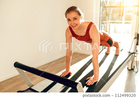 Slim woman wearing sports attire having anaerobic exercise on reformer Slim woman wearing sports attire having anaerobic exercise on reformer 112499320