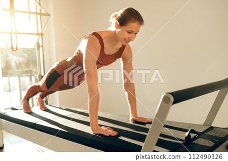 Slim woman wearing sports attire having anaerobic exercise on reformer Slim woman wearing sports attire having anaerobic exercise on reformer 112499326