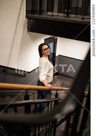 Satisfied woman wearing casual clothes and glasses walking on stairs in hotel Satisfied woman wearing casual clothes and glasses walking on stairs in hotel 112499460