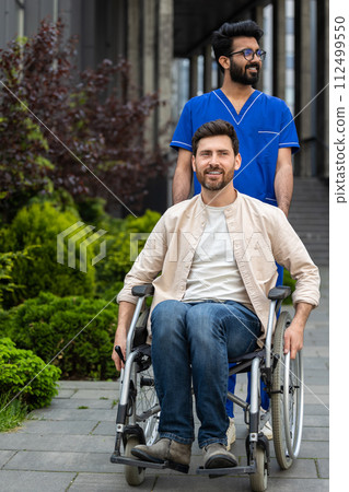 Disabled man sitting in a wheelchair, male nurse rolling it 112499550