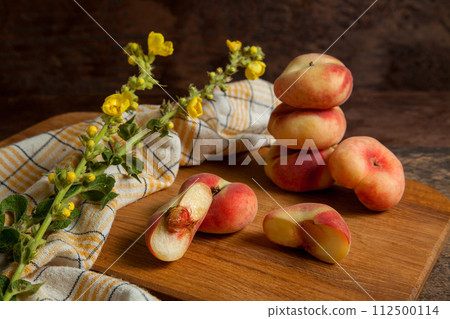 Cutting board with halves and several whole saturn peaches or flat peaches on wooden background with yellow kitchen towel.. Cutting board with halves and several whole saturn peaches or flat peaches on wooden background with yellow kitchen towel.. 112500114